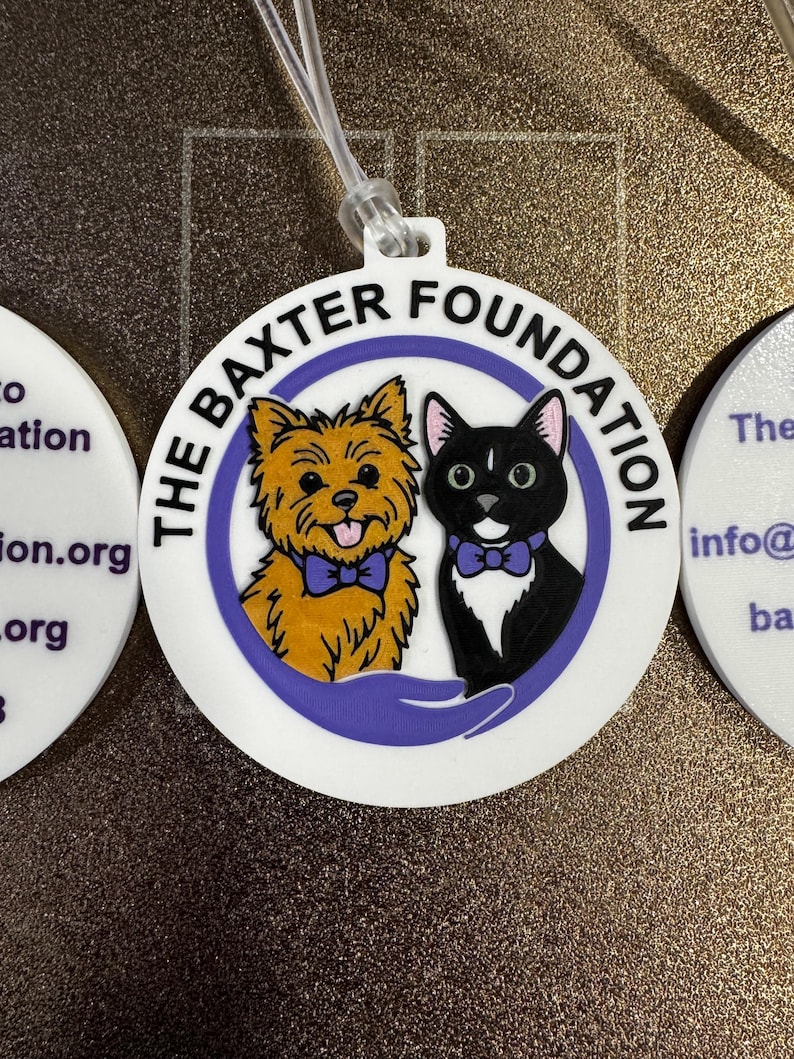 May include: White, round tag featuring a cartoon dog and cat wearing bow ties, with the text "THE BAXTER FOUNDATION" in a purple ring. A clear plastic loop is attached at the top. The tag is likely for a pet.