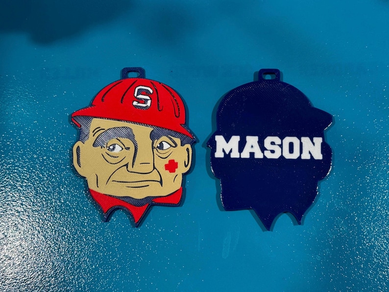 May include: Two decorative ornaments. One features a cartoon face wearing a red hard hat with the letter "S". The other is a blue silhouette with the name "MASON" in white block letters. Both have a small hole at the top.