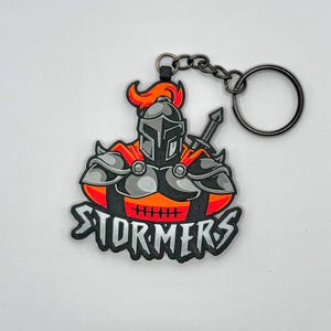 May include: A black and silver Stormers keychain featuring a knight in armor with an orange plume and cape. The keychain has a black chain and a silver key ring. The word "STORMERS" is written in silver.