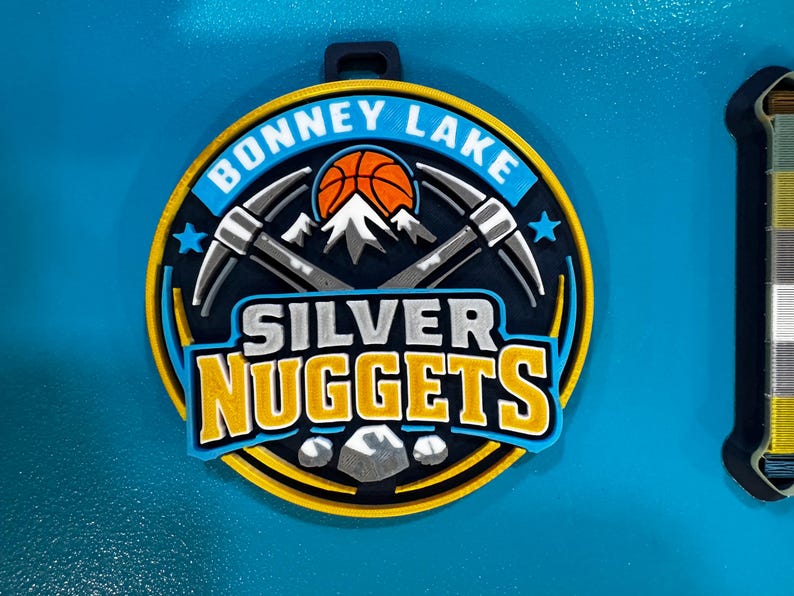 May include: A circular, multi-layered emblem with the text "BONNEY LAKE" and "SILVER NUGGETS". The design includes a basketball, crossed pickaxes, and a mountain range. The color scheme is blue, yellow, orange, gray, and white.
