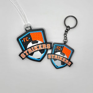 May include: Two soccer-themed keychains and luggage tags. Each item features a black shield with orange and blue accents, a soccer ball graphic, and the word "STRIKERS". One has a keychain ring, the other a clear plastic strap.