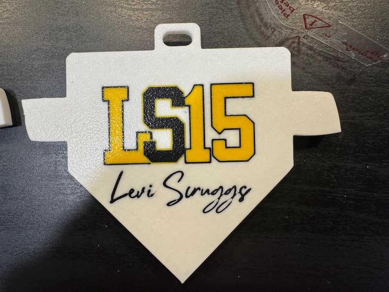May include: A white baseball home plate-shaped tag with the text "LS15" in yellow and black, and the name "Levi Scruggs" in black script. The tag has a small hole at the top for hanging.