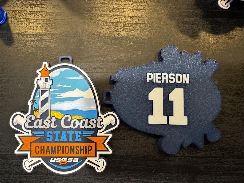 May include: Two sports-themed medals. One features a lighthouse, the text "East Coast State Championship", and crossed baseball bats. The other is navy blue with the name "Pierson" and the number "11" in white.