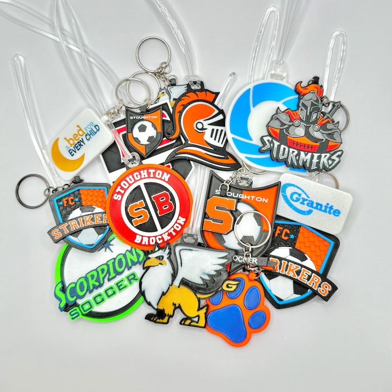 May include: A collection of colorful keychains with various soccer team logos and mascots. Designs include soccer balls, eagles, and team names like "STORMERS" and "SCORPIONS SOCCER". Each keychain has a clear plastic loop and a metal keyring.