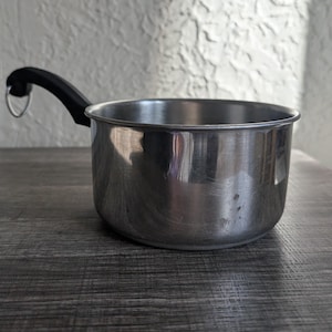 May include: A small, stainless steel saucepan with a black handle and a silver ring for hanging. The pot is round with a slightly flared rim and sits on a dark wooden surface. The handle is attached to the side of the pot.