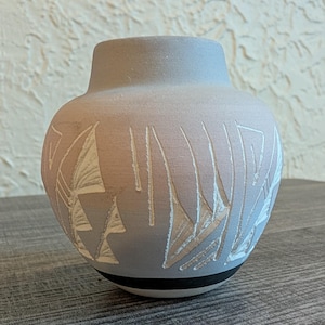 Peut inclure: Un vase en céramique grise avec un corps arrondi et un col étroit. Le vase présente un motif géométrique blanc sculpté. Une bande noire entoure la base. Le vase est exposé sur une surface en bois.