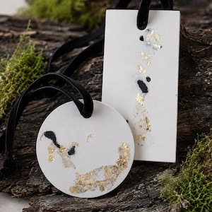 May include: Two white hanging ornaments, one circular and one rectangular, featuring black accents and gold leaf flakes. Each ornament has a hole at the top threaded with a black velvet ribbon for hanging, creating a contrast between the white surface.