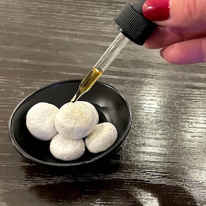 May include: A black, oval-shaped bowl filled with several white, round objects. A clear dropper dispenses a golden liquid onto the objects. The dropper has a black rubber top. The objects appear to have a textured surface.