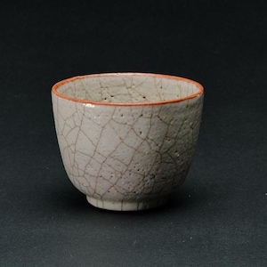 Może przedstawiać: Mała, kremowobiała ceramiczna miska z pękniętym wzorem szkliwa. Obręcz jest w kolorze czerwonopomarańczowym. Miska ma zaokrąglony kształt i stoi na małej, nieszkliwionej podstawie. Tło jest jednolicie czarne.