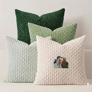May include: A set of four square throw pillows with a textured, ribbed corduroy-style fabric. The pillows are arranged in a stack, featuring shades of dark forest green, sage green, light mint green, and cream, with a small illustration of a couple.