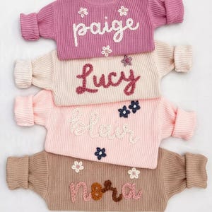 May include: Four long-sleeved knit sweaters stacked vertically. The top is mauve with white text "paige" and small white flowers. The second is cream with dark pink text "Lucy" and a small flower. The third is light pink with white text "blair" and dark blue flowers. The bottom is tan with pink and brown text "nora" and white flowers.