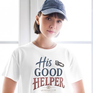May include: A white short-sleeved t-shirt featuring the blue text His, the blue text GOOD, and the red text HELPER. A graphic of a diamond ring is positioned next to the word His. Below the text is a small ring icon and the text GENESIS 2:18.