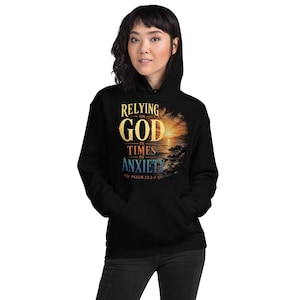 Może przedstawiać: Czarna bluza z kapturem z nadrukiem graficznym zawierającym tekst RELYING ON GOD IN TIMES OF ANXIETY oraz PSALM 23:2-3. Wzór przedstawia zachód słońca nad taflą wody ze skalistymi brzegami, utrzymany w ciepłych złotych i niebieskich odcieniach.