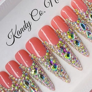 May include: A set of Kandy Co. Nails, featuring long, almond-shaped press-on nails. Each nail is painted a soft coral color with a bedazzled design of iridescent rhinestones along the tips. The nails are arranged in a white box.