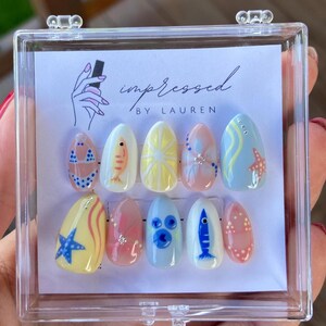 Pu&ograve; includere: Un set di dieci unghie finte ovali in una custodia di plastica trasparente. Le unghie sono dipinte con vari disegni a tema spiaggia, tra cui un bikini, un pesce, un sole, una stella marina e pois. Il testo "impressed by Lauren" &egrave; visibile.