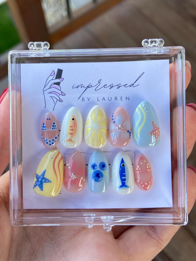 Pu&ograve; includere: Un set di dieci unghie finte ovali in una custodia di plastica trasparente. Le unghie sono dipinte con vari disegni a tema spiaggia, tra cui un bikini, un pesce, un sole, una stella marina e pois. Il testo "impressed by Lauren" &egrave; visibile.