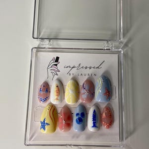 Pu&ograve; includere: Un set di dieci unghie finte in una custodia di plastica trasparente. Ogni unghia &egrave; dipinta con un design diverso, tra cui pesci, stelle marine e motivi astratti nei toni del blu, giallo, rosso e bianco. Il testo "impressed by Lauren" &egrave; visibile.