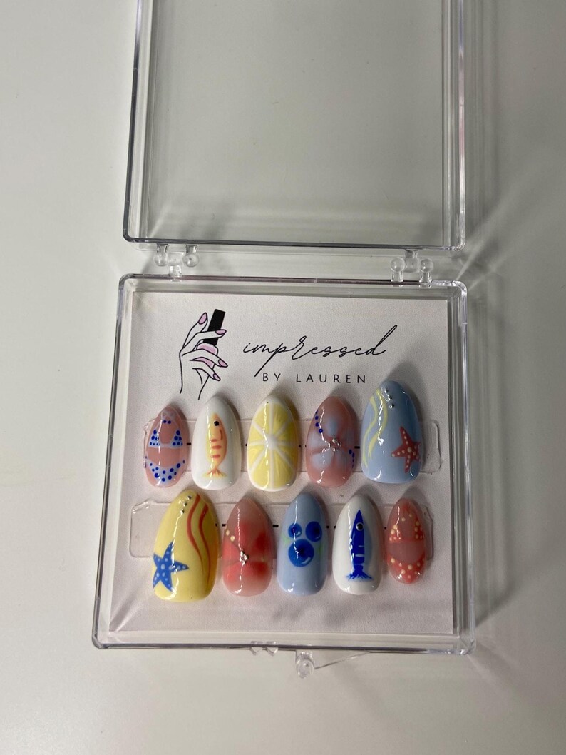 Pu&ograve; includere: Un set di dieci unghie finte in una custodia di plastica trasparente. Ogni unghia &egrave; dipinta con un design diverso, tra cui pesci, stelle marine e motivi astratti nei toni del blu, giallo, rosso e bianco. Il testo "impressed by Lauren" &egrave; visibile.