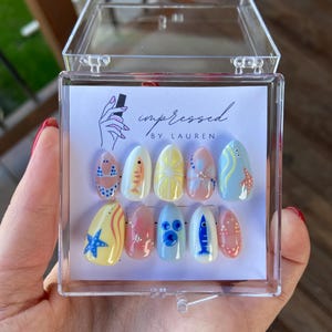 Pu&ograve; includere: Un set di dieci unghie finte a tema estivo, con disegni di pesci, stelle marine e motivi bikini. Le unghie sono esposte in una scatola di plastica trasparente. Il testo "impressed by Lauren" &egrave; visibile.