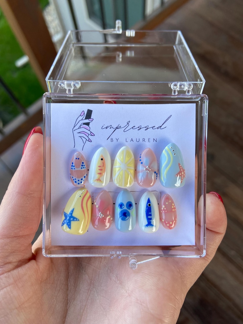 Pu&ograve; includere: Un set di dieci unghie finte a tema estivo, con disegni di pesci, stelle marine e motivi bikini. Le unghie sono esposte in una scatola di plastica trasparente. Il testo "impressed by Lauren" &egrave; visibile.