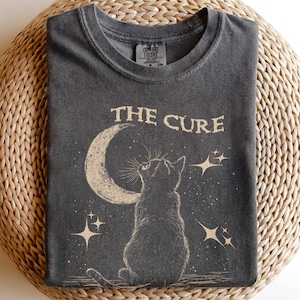May include: A charcoal gray t-shirt featuring a cream-colored graphic of a cat sitting and looking up at a crescent moon and stars. The text THE CURE is printed above the cat. The shirt has a crew neckline and a Comfort Colors brand label.
