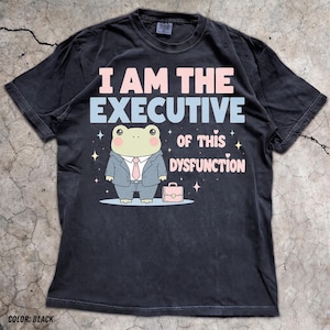 Op de afbeelding: Een zwart t-shirt met korte mouwen en een afbeelding van een cartoonkikker in een grijs pak met een roze das. De tekst luidt I AM THE EXECUTIVE OF THIS DYSFUNCTION. Het ontwerp bevat een kleine roze aktetas en diverse kleine sterren.