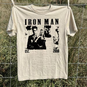 May include: Cream-coloured t-shirt with a black graphic that reads "IRON MAN" above a collage of images. The graphic includes the text "EST. 2008". The t-shirt is hanging on a clothesline against a backdrop of green grass and a chain-link fence.