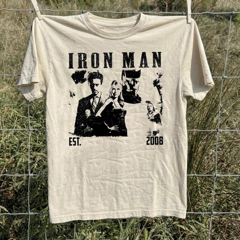 May include: Cream-coloured t-shirt with a black graphic that reads "IRON MAN" above a collage of images. The graphic includes the text "EST. 2008". The t-shirt is hanging on a clothesline against a backdrop of green grass and a chain-link fence.