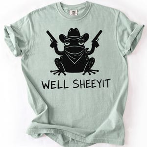 Peut inclure: Un t-shirt à manches courtes vert sauge clair avec un motif noir représentant une grenouille portant un chapeau de cowboy et un bandana, tenant deux pistolets. Sous la grenouille, le texte WELL SHEEYIT est imprimé en police noire.