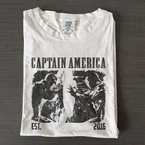 May include: White t-shirt with a black graphic of Captain America and the text "CAPTAIN AMERICA" above the graphic. The shirt also has the text "EST. 2016". The shirt is folded neatly on a dark surface.