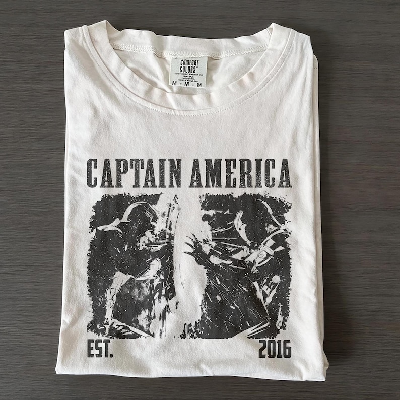 May include: White t-shirt with a black graphic of Captain America and the text "CAPTAIN AMERICA" above the graphic. The shirt also has the text "EST. 2016". The shirt is folded neatly on a dark surface.