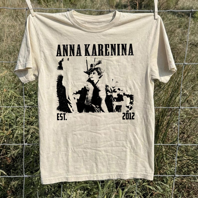 Puede incluir: Camiseta color crema con el texto "ANNA KARENINA" sobre un gr&aacute;fico en blanco y negro de una mujer con sombrero. El texto "EST. 2012" est&aacute; impreso debajo. La camiseta est&aacute; colgada en un tendedero.