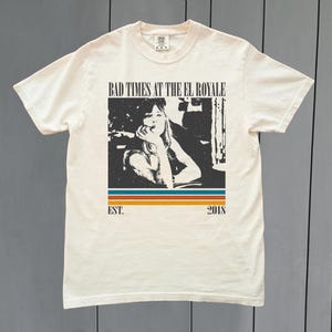 May include: Cream-colored t-shirt featuring a black and white graphic with the text "BAD TIMES AT THE EL ROYALE" above a picture of a woman. Below the image are the words "EST. 2018" and a horizontal stripe design.