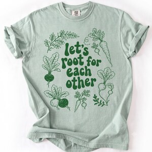 Peut inclure: Un t-shirt à manches courtes vert sauge clair orné d'illustrations au trait vert foncé représentant divers légumes-racines entourant le texte "let's root for each other" imprimé dans une police arrondie et grasse vert foncé sur la poitrine.
