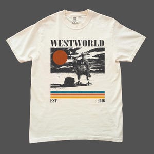 May include: Off-white t-shirt with a graphic print featuring the word "WESTWORLD" above a desert scene with a bird and a setting sun. Below the image are horizontal stripes and the text "EST. 2016".