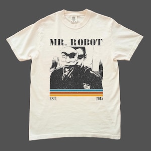 Könnte beinhalten: Cremefarbenes T-Shirt mit einer schwarz-weißen Grafik von zwei Figuren, eine davon maskiert. Die Worte "MR. ROBOT" stehen über der Grafik, darunter "EST. 2015". Ein horizontaler Streifen in Blau, Orange und Braun befindet sich darunter.