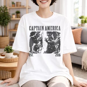 May include: White t-shirt featuring a black and white graphic of Captain America. The shirt has the words "CAPTAIN AMERICA" printed across the top, with "EST. 2016" below the graphic. The shirt is a casual, oversized fit.