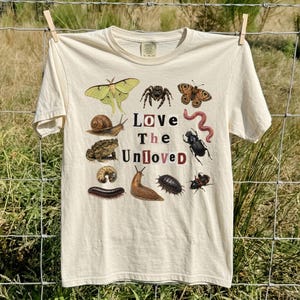 May include: A cream-coloured short-sleeve t-shirt featuring a graphic print of various insects and small creatures, including a moth, spider, snail, toad, and worm. The text "Love The Unloved" is printed in the centre using a ransom note style font.