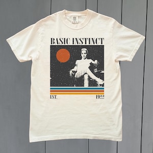 Puede incluir: Camiseta color crema con un diseño gráfico retro. El diseño incluye el texto "BASIC INSTINCT" sobre una imagen de una mujer. Debajo, se lee "EST. 1992". El diseño presenta un círculo rojo y rayas horizontales.