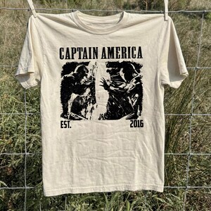 May include: Cream-colored t-shirt featuring a black and white graphic of Captain America. The text "CAPTAIN AMERICA" is printed above the graphic, with "EST. 2016" below. The shirt is hanging on a clothesline against a backdrop of green grass and a wire fence.
