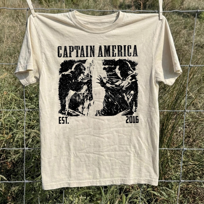 May include: Cream-colored t-shirt featuring a black and white graphic of Captain America. The text "CAPTAIN AMERICA" is printed above the graphic, with "EST. 2016" below. The shirt is hanging on a clothesline against a backdrop of green grass and a wire fence.