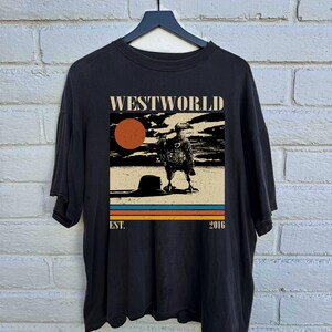 May include: Black t-shirt featuring a graphic from the TV show "Westworld." The design includes the show's title, a vulture, a setting sun, and the text "EST. 2016" in a retro style. The shirt is hanging on a black hanger.