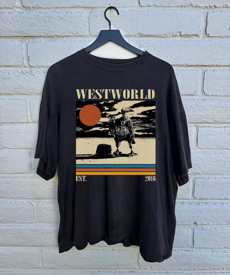 May include: Black t-shirt featuring a graphic from the TV show "Westworld." The design includes the show's title, a vulture, a setting sun, and the text "EST. 2016" in a retro style. The shirt is hanging on a black hanger.