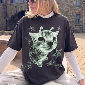 Könnte beinhalten: Ein anthrazitfarbenes Kurzarm-T-Shirt mit einer hellgrünen Grafik einer Katze, die zwei Antennen auf dem Kopf trägt und von mehreren Sternen umgeben ist. Das T-Shirt wird über einem weißen Langarmshirt und einer hellen Hose getragen.