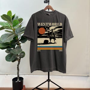 May include: Dark gray t-shirt with a graphic from the show Westworld. The graphic features a vulture, a setting sun, and the text "WESTWORLD" above the image and "EST. 2016" below. The shirt is hanging on a wooden hanger.