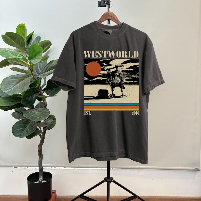 May include: Dark gray t-shirt with a graphic from the show Westworld. The graphic features a vulture, a setting sun, and the text "WESTWORLD" above the image and "EST. 2016" below. The shirt is hanging on a wooden hanger.