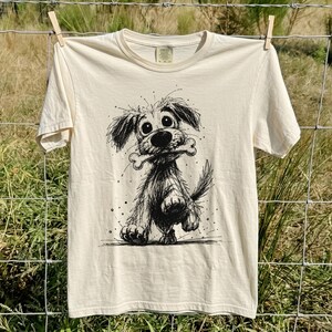 Op de afbeelding: Een crèmekleurig T-shirt met korte mouwen en een zwarte inkttekening van een ruige hond met grote ogen die een bot in zijn bek houdt. De afbeelding staat centraal op de borst en het shirt heeft een ronde hals en een zichtbaar neklabel.