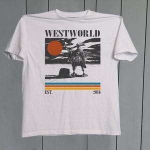 May include: White t-shirt with a graphic design featuring the word "WESTWORLD" above an illustration of a bird and a sunset. Below the image are the words "EST. 2016" and a series of horizontal stripes.