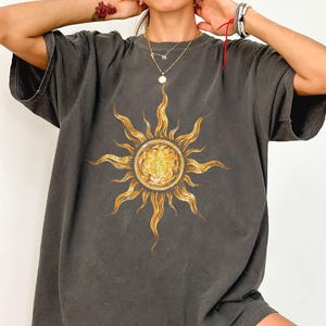 May include: A charcoal gray short-sleeve t-shirt featuring a large, centered graphic of a golden yellow sun with wavy, elongated rays. The shirt has a crew neckline and a relaxed fit, showcasing a detailed illustration of the sun's surface.