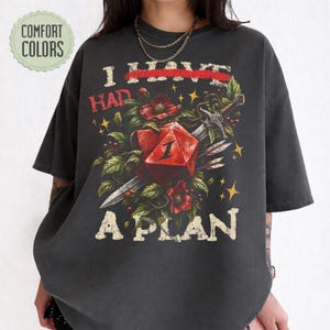 Könnte beinhalten: Ein anthrazitfarbenes T-Shirt mit einem Grafikdruck eines roten zwanzigseitigen Würfels, der von einem Schwert durchbohrt wird, umgeben von roten Blumen und grünen Blättern. Der Text lautet I HAD A PLAN mit einem roten Strich durch HAD. Comfort Colors Logo.